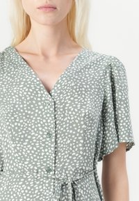 Kobieta w lekkiej zielonej sukience w białe nieregularne kropki, z dekoltem w serek, guzikami z przodu, krótkimi rękawami i przewiązanym paskiem w talii.