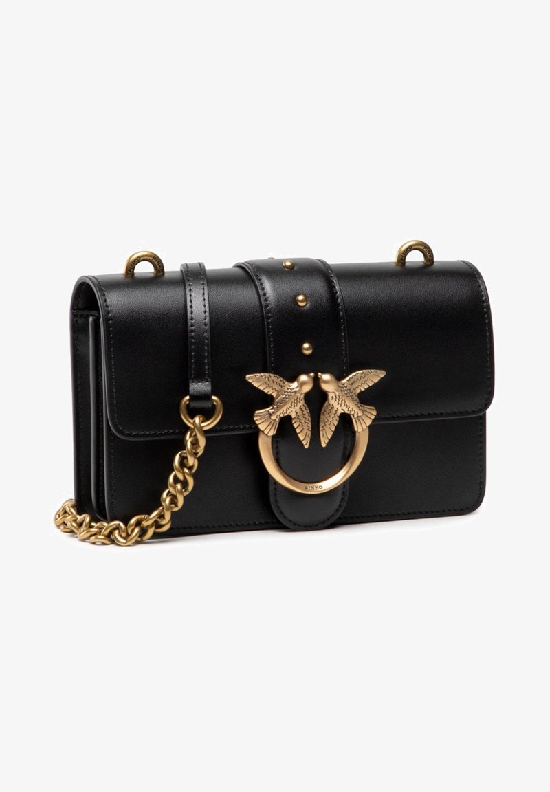 Bolsa de mão em couro preto com fecho de aba, alça de corrente dourada e um anel decorativo com detalhes de dois pássaros na parte da frente.