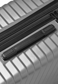 Close-up van een zilveren harde koffer met horizontale ribbels, een zwarte plastic zijhandgreep en een dubbele rits met trekbandjes.