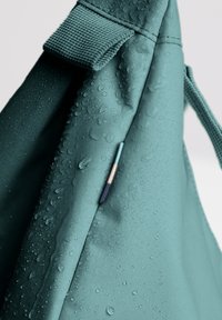 Turkos vattentätt ryggsäck gjord av texturerad tyg, med droppar på ytan och en liten metallisk detalj nära sömmen.