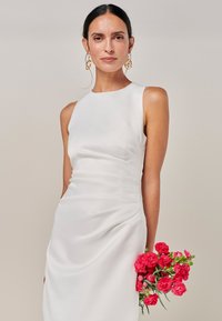 Vit ärmlös klänning i slätt tyg med rynkad detalj vid midjan. Modellen håller en bukett rosa blommor.