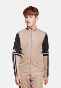 Beige und schwarze Zip-Jacke mit langen Ärmeln, die drei Streifen an den Ärmeln und ein Logo auf der Brust hat. Glattes Material, athletische Passform.