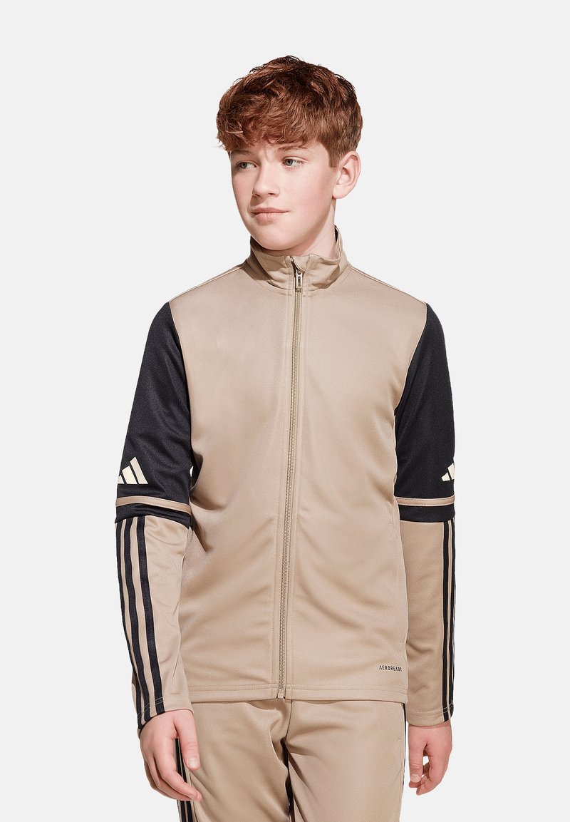 Beige und schwarze Zip-Jacke mit langen Ärmeln, die drei Streifen an den Ärmeln und ein Logo auf der Brust hat. Glattes Material, athletische Passform.