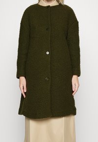 Cappotto lungo verde scuro con trama a tre bottoni indossato sopra una camicia beige con colletto e una gonna crema, su una persona in piedi.