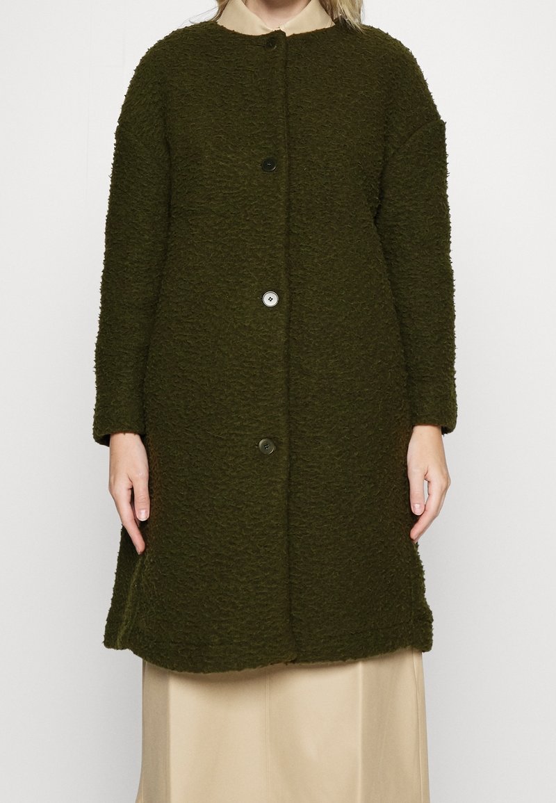 Cappotto lungo verde scuro con trama a tre bottoni indossato sopra una camicia beige con colletto e una gonna crema, su una persona in piedi.