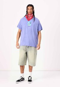 Camiseta de gran tamaño en color púrpura claro con un parche gráfico, combinada con pantalones cortos de mezclilla descoloridos hasta las rodillas, bandana roja y zapatillas negras.