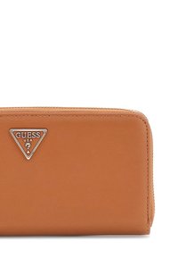 Tan leather wallet with a textured finish, featuring a silver triangular Guess logo and a zippered closure along the top edge.