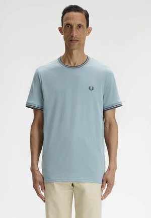 Hombre con camiseta azul claro de manga corta con ribete a rayas en el cuello y las mangas, pantalones beige, de pie frente a un fondo liso.