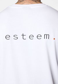 Weißes Baumwoll-T-Shirt mit dem Wort "esteem" in schwarzem Stick auf der Rückseite, ergänzt durch einen kleinen roten Punkt. Einfaches und minimalistisches Design.