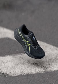 Svart och gul Asics löparsko svävande i luften ovanför asfalt med vit vägmarkering och små gruskorn.