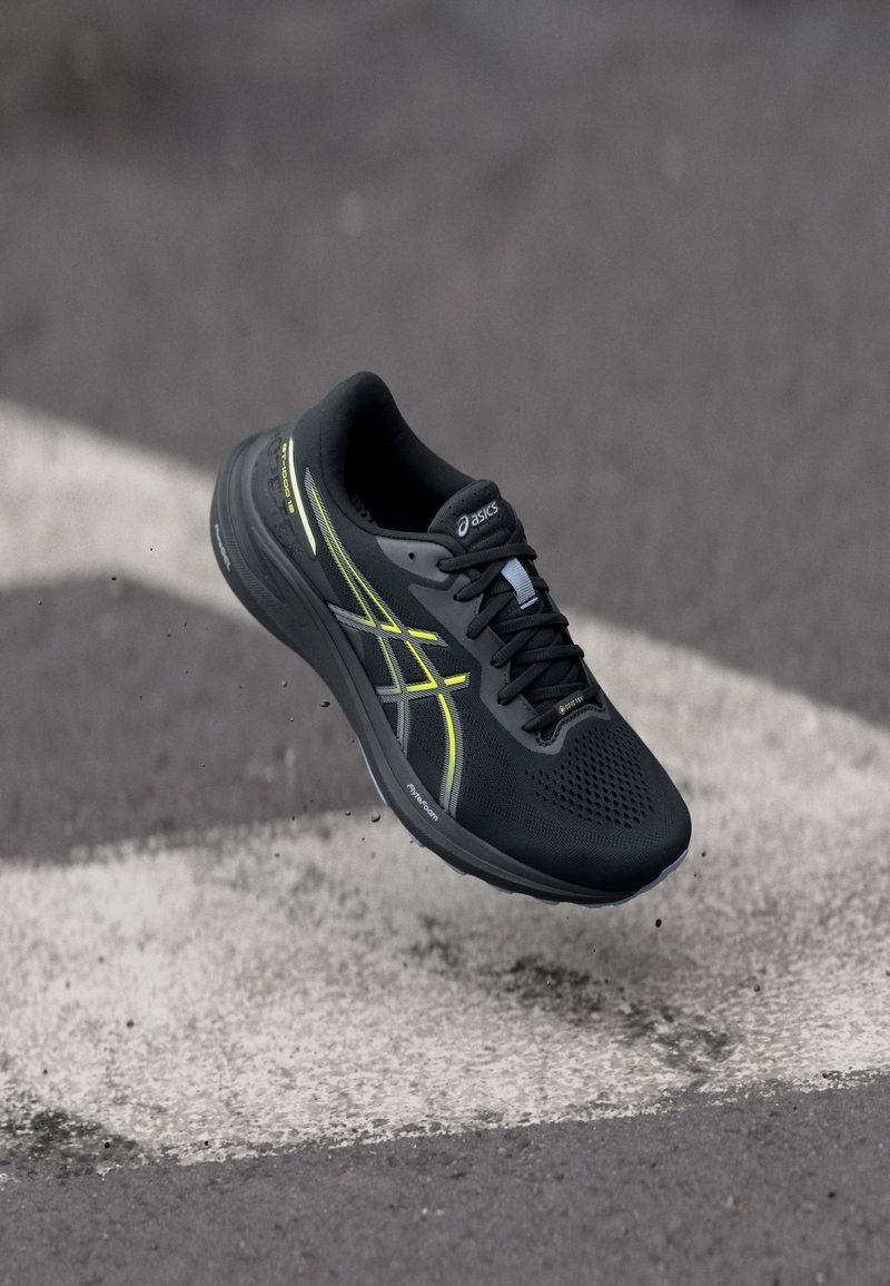 Sort og gul Asics løbesko svævende i luften over asfalt med hvid vejmarkering og små gruspartikler.