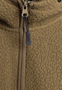 Giacca in pile marrone con superficie testurizzata, dotata di zip in metallo e tirante in corda nera. Include dettagli marroni lungo la zip.