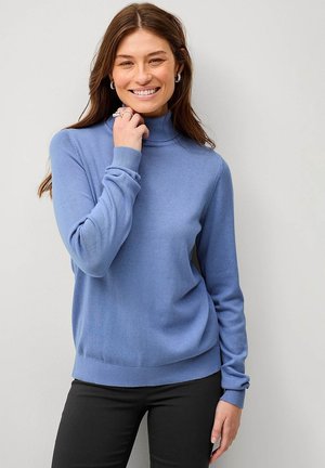 Lichtblauwe coltrui van zachte brei met ribbelmanchetten en -zoom, gedragen met zwarte broeken, model sorrend en haar gezicht aanrakend.