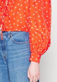 Blouse longue à manches longues de couleur orange vif avec de petits motifs floraux blancs, poignets bouffants, détail noué sur le côté, associée à un jean en denim bleu.