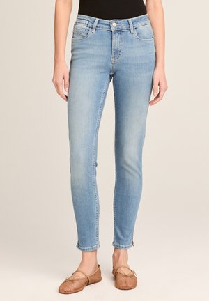 Frau trägt hellblaue Skinny-Jeans und hellbraune gewebte Slipper und steht vor einem schlichten beigen Hintergrund.