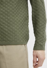 Olijfgroen gebreid vest met een gestructureerd geblokt patroon, ribbels aan de mouwen en zoom. Gecombineerd met lichtbeige broek.