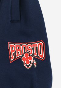 Granatowy sweatshirt z wyraźnym czerwono-białym logo "PROSTO" oraz czerwonym wzorem koniczyny. Materiał wydaje się miękki i gładki w dotyku.
