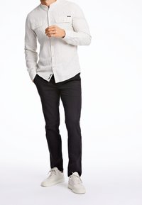 Man wearing light beige button-up shirt, black pants, and white sneakers, standing with one hand near chest and the other in pocket.