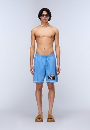 Uomo senza maglietta che indossa pantaloncini da nuoto azzurri con logo nero, sandali marroni a ciabatta, collana di conchiglie e occhiali da sole con lenti viola, in piedi di fronte.