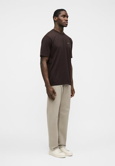 Νέος μαύρος άνδρας που στέκεται, φοράει σκουρό καφέ μπλουζάκι, μπεζ παντελόνι και λευκά αθλητικά παπούτσια μπροστά σε απλό λευκό φόντο.