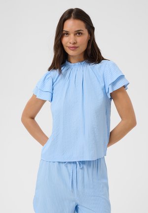 Femme aux cheveux bruns portant un haut bleu clair à manches courtes et un pantalon à cordon assorti, debout les mains sur les hanches devant un fond uni.