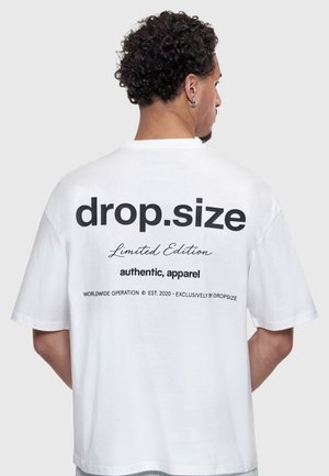 Bărbat cu păr creț purtând tricou alb cu text negru „drop.size Limited Edition authentic, apparel”, văzut din spate pe fundal gri.