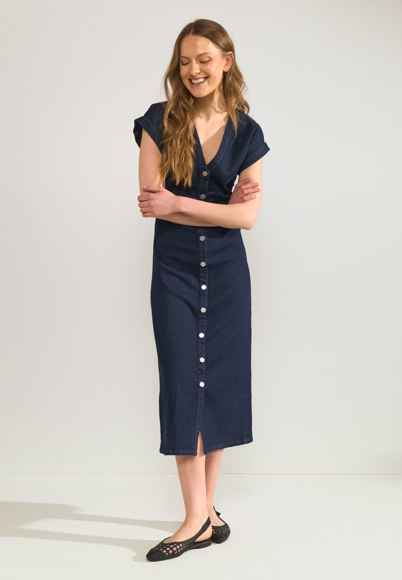 Jeune femme aux longs cheveux portant une robe midi bleu foncé boutonnée et des ballerines tissées noires, debout les bras croisés et souriante.