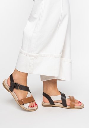 Pieds portant des sandales en cuir marron, noir et blanc avec des brides à la cheville, associées à un pantalon blanc à jambes larges.