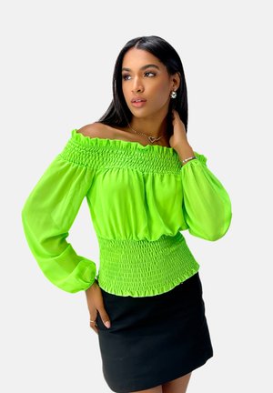 Groene, off-the-shoulder blouse met smock-borst en lange mouwen. Doorzichtig materiaal met een lichte glans, gedragen over een zwarte, aansluitende rok.