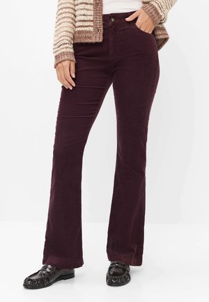 Kvinde iført mørkelilla kordfløjlsbukser, stribet beige cardigan og sorte loafers, stående med den ene hånd i lommen.