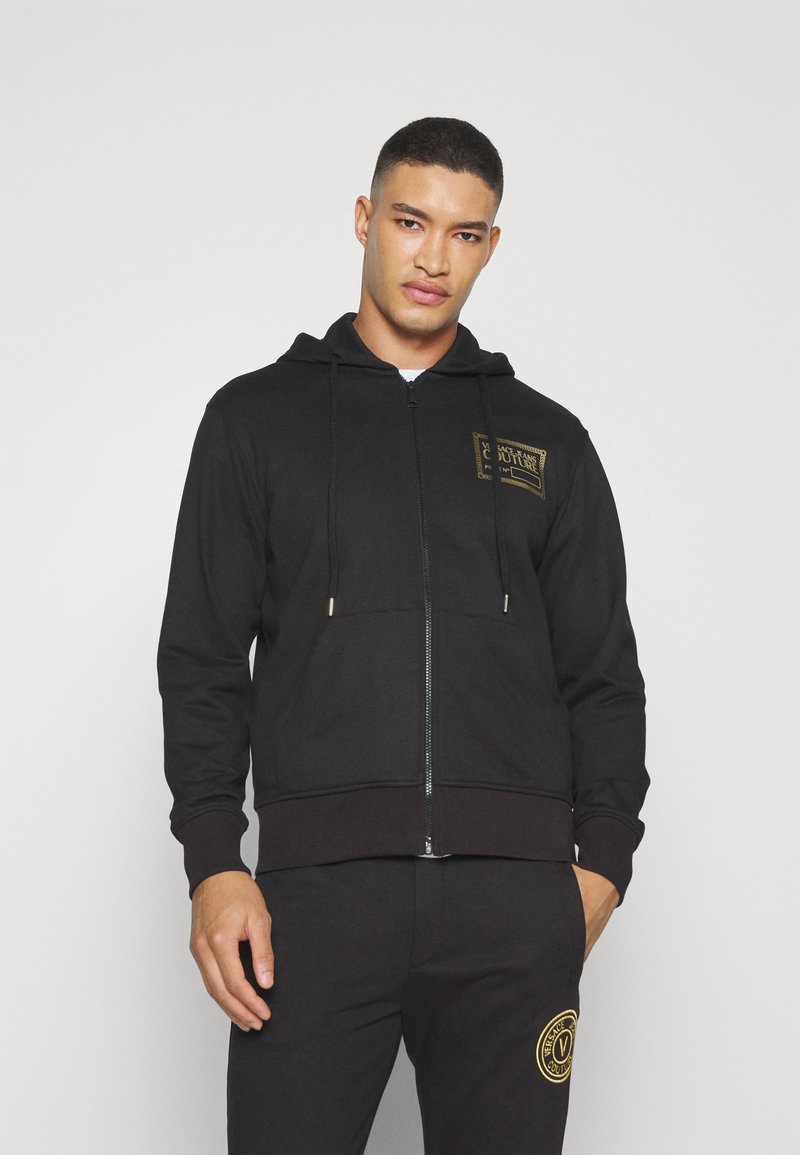 Zwarte zip-up hoodie met een vozak pocket, verstelbare trekkoordcapuchon en een gouden logo patch op de borst. Zachte stof en geribde mouwen.