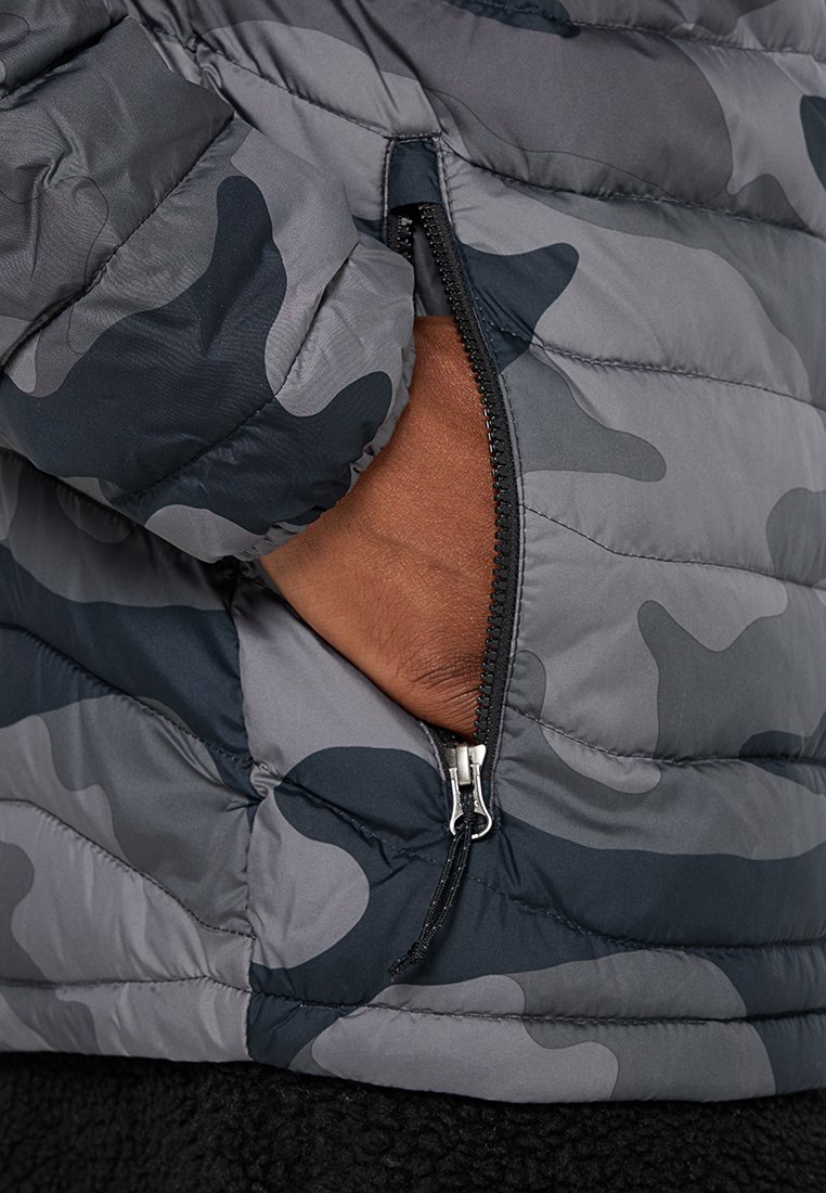 Chaqueta tipo abrigo inflable con un patrón de camuflaje en gris y azul marino, secciones acolchadas, bolsillo con cremallera y textura exterior suave.