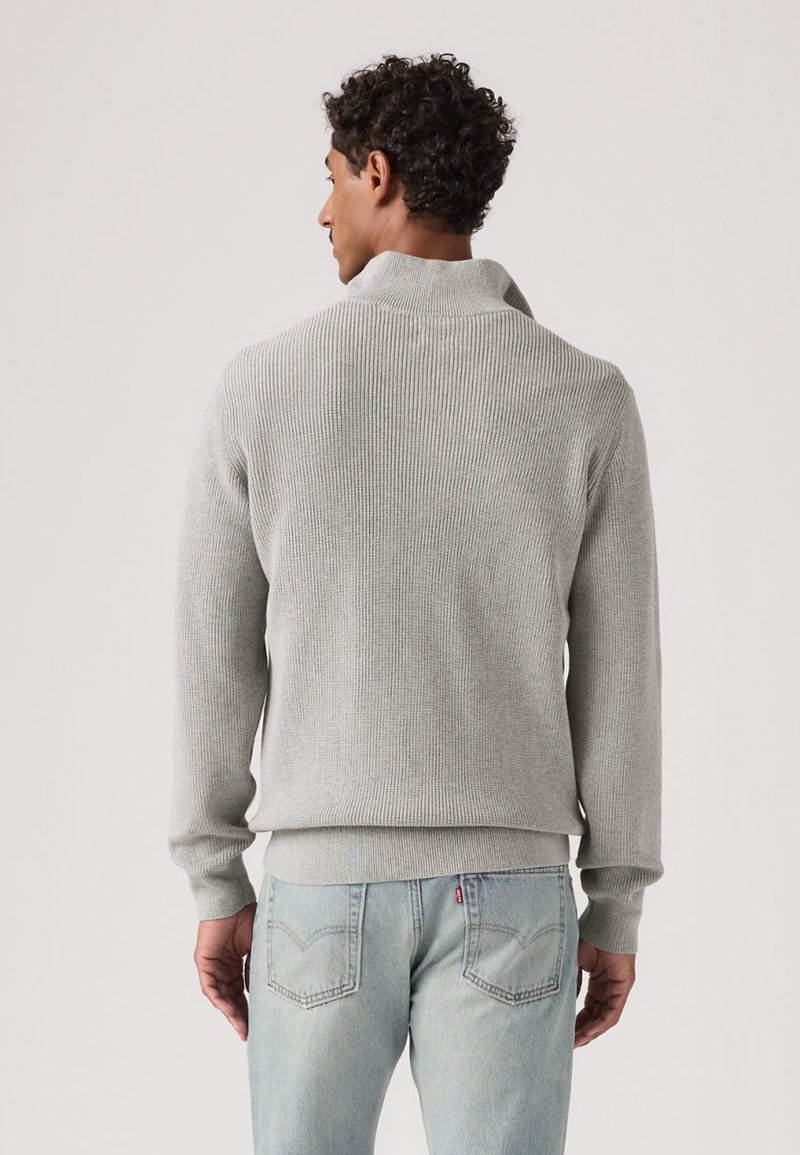 Pull en maille côtelée gris avec un col montant, cintré à la taille et manches longues, assorti à un jean bleu clair à coupe décontractée.
