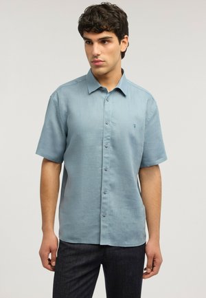 Ung mand stående iført en lyseblå kortærmet skjorte med knapper og mørke bukser, kigger let til siden.