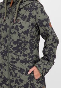 Hand van een vrouw in de zak van een groen en zwart bloemenpatroon, met ritssluiting en capuchon met trekkoord.