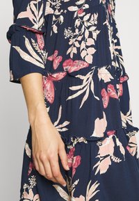 Marinblå blommig klänning med rosa och krämfärgade blommor, lagerdesign, volangärmar och en strukturerad tygkvalitet. Visar en hand som vilar på plagget.