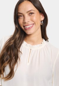 Krämfärgad blus med subtil prickig textur, volangkrage och liten nyckelhålsdetalj. Modellen bär hoopsörhängen.