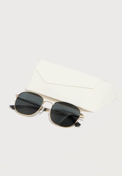 Ορθογώνια γυαλιά ηλίου με μαύρους φακούς και λεπτό χρυσό πλαίσιο τοποθετημένα μπροστά από μια λευκή γεωμετρική θήκη γυαλιών σε λευκό φόντο.