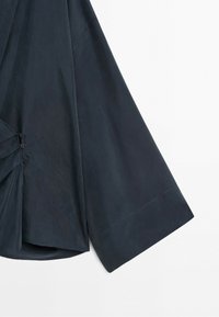 Detail tmavě modré košile s dlouhým rukávem, širokými manžetami a na bočním švu zapnutými záhyby.