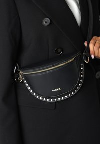 Svart läder Mexx bälte väska med gulddragkedja och nitar som bärs crossbody över en svart kappa, hand som håller i remmen med ring och diamantring synlig.