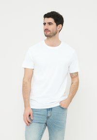 Άνδρας που φοράει απλό λευκό κοντομάνικο μπλουζάκι και ανοιχτό μπλε τζιν παντελόνι, στέκεται με το ένα χέρι στην τσέπη, κοιτώντας προς τα πλάγια.