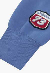 Ανοιχτό μπλε μανίκι φούτερ με ριγέ μανσέτα και κεντητή ετικέτα με το "Original 73" σε κόκκινο και άσπρο χρώμα σε φόντο ασπίδας.