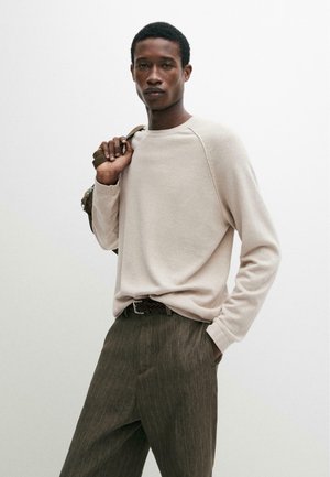 Mand iført beige langærmet skjorte og olivengrønne bukser med striber, der holder rygsætsrem over skulderen og poserer mod en ensfarvet hvid baggrund.