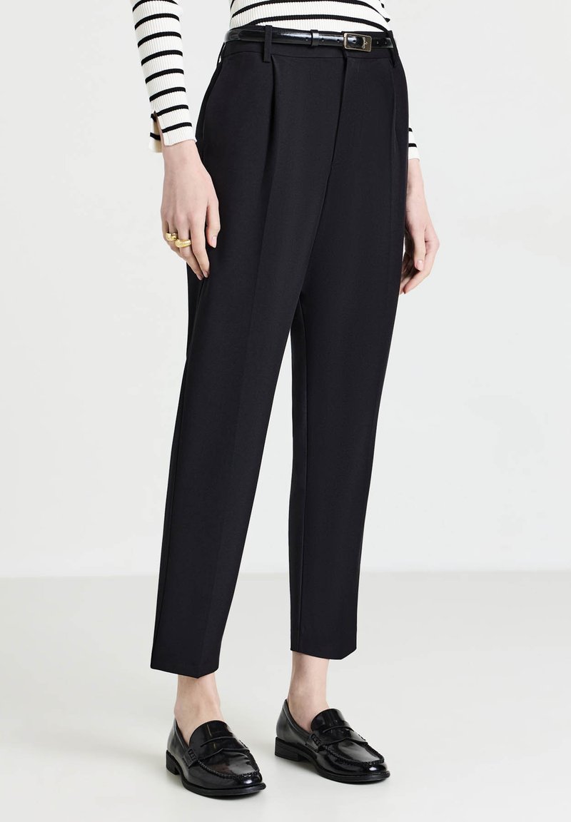 Pantalons noirs tailleur au toucher lisse, dotés d'une taille haute, de plis et d'une coupe droite. Portés avec une ceinture noire et des mocassins.