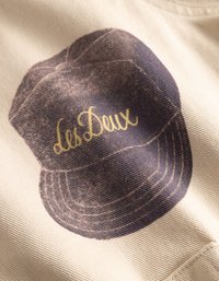 Το μπεζ ύφασμα διαθέτει ένα σκούρο μπλε καπέλο με εκτύπωση και υφή, που αικοσμείται από το χρυσό κείμενο "Les Deux."