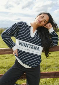 Camicia a maniche lunghe a righe blu navy e bianca con colletto, con la scritta "MONTANA USA", abbinata a jeans scuri.