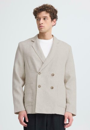 Ung mand med krøllet hår iført beige dobbeltradet blazer over hvid skjorte, stående foran en ensfarvet lysegrå baggrund.