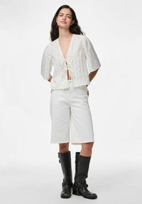 Bílá krátkosrstá blůza s výšivkovým vzorem a detaily na zavazování v přední části, v kombinaci s bílými šortkami do kolen a černými vysokými botami.