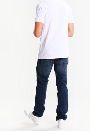 T-shirt bianca a maniche corte e jeans in denim blu scuro; i jeans hanno una vestibilità standard e un design a cinque tasche, abbinati a sneaker bianche.