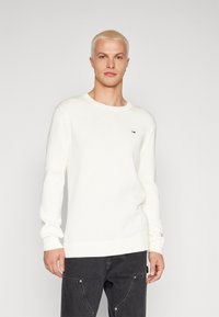Suéter blanco de punto con cuello redondo, puños y dobladillo acanalados, que presenta un pequeño logo en el pecho. El modelo lleva pantalones oscuros.